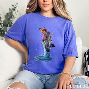 May include: A blue t-shirt with a graphic of characters from the film Zootropolis. The design includes a fox, a rabbit, and a snake. The shirt is a solid blue colour and has the words "FLO BLUE" printed at the bottom.