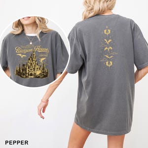 May include: Pepper-colored t-shirt with a graphic on the front featuring a castle, trees, and the text "Riorrson House". The back of the shirt has a gold dragon design. The shirt is a comfortable, oversized fit.