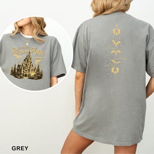 May include: A grey t-shirt featuring a castle graphic with the text "Peterson's House" in gold on the front. The back of the shirt has a gold design of dragons and clouds. The shirt is a short-sleeved, oversized fit.