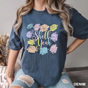 May include: A denim blue t-shirt with a colorful seashell design and the words "Shell Yeah" in a script font. The seashells are in various colors, including yellow, pink, blue, and green.