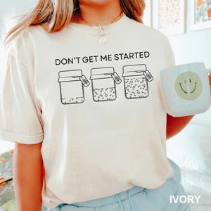 May include: Ivory t-shirt featuring the phrase "DON'T GET ME STARTED" above three jars with tags. The jars contain different amounts of dots. A white mug with a smiley face is visible on the right.