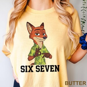 Puede incluir: Camiseta amarilla clara con un personaje de zorro de dibujos animados con una camisa hawaiana verde, haciendo el gesto de "ok". El texto "SIX SEVEN" est&aacute; impreso en letras negras debajo.