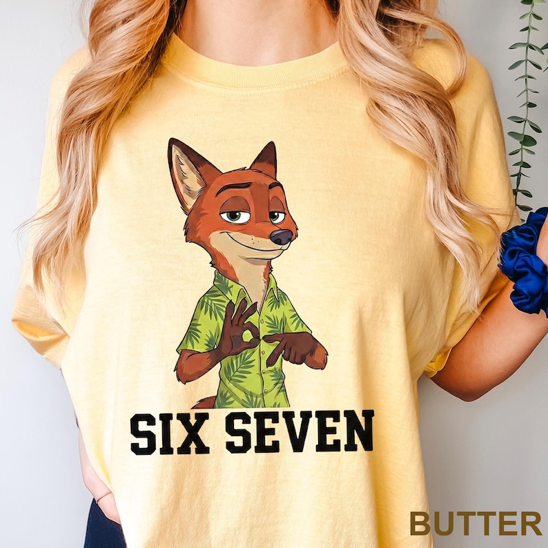 Puede incluir: Camiseta amarilla clara con un personaje de zorro de dibujos animados con una camisa hawaiana verde, haciendo el gesto de "ok". El texto "SIX SEVEN" est&aacute; impreso en letras negras debajo.