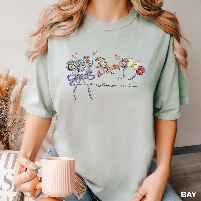 Puede incluir: Camiseta verde claro con dise&ntilde;os de personajes de dibujos animados y el texto "You can be anything you wish to be." El dise&ntilde;o incluye una placa de polic&iacute;a, una zanahoria, un zorro, una rosquilla y una frambuesa.