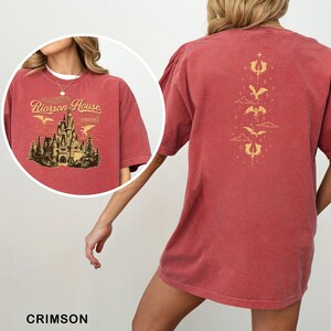 May include: Crimson-colored t-shirt featuring a gold illustration of a castle with the text "Riorison House" and "Aretian". The back of the shirt has a gold design of dragons and clouds. The shirt is a comfortable, oversized fit.