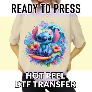 May include: Beige t-shirt featuring a colorful graphic of a blue cartoon character riding a surfboard in front of a wave. The design includes pink flowers and the text "READY TO PRESS" and "HOT PEEL DTF TRANSFER".