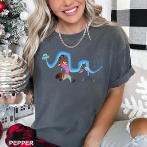 Może przedstawiać: Ciemnoszara koszulka z kresk&oacute;wkową ilustracją lisa i kr&oacute;lika biegnących, z niebieskim wzorem przypominającym węża nad nimi. Koszulka ma podwinięte rękawy i luźny kr&oacute;j. Słowo "PEPPER" jest widoczne na dole.