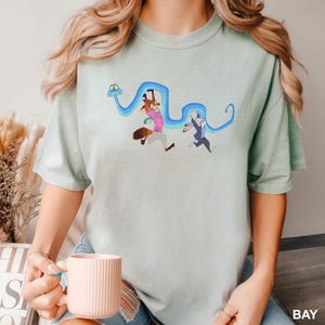 Może przedstawiać: Jasnozielona koszulka z kresk&oacute;wkową ilustracją lisa i kr&oacute;lika biegnących z niebieskim wężem. Lis ma na sobie r&oacute;żową koszulę, a kr&oacute;lik niebieski garnitur. Słowo "BAY" jest wydrukowane na dole po prawej stronie na biało.