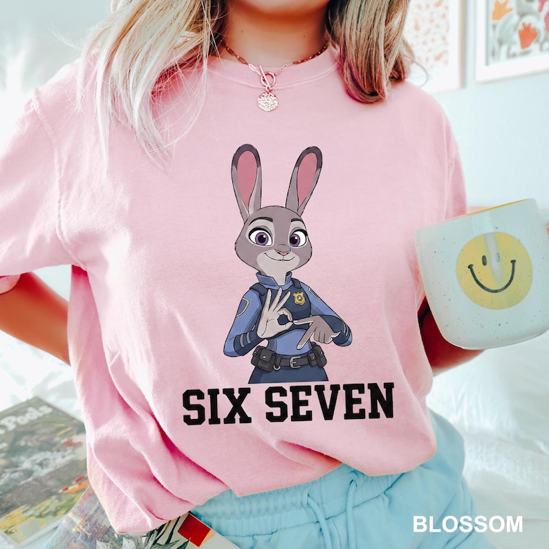 Puede incluir: Sudadera rosa claro con una imagen de dibujos animados de un conejo polic&iacute;a de Zootopia, con el texto "SIX SEVEN" en letras negras. La palabra "BLOSSOM" est&aacute; impresa en blanco en la parte inferior.