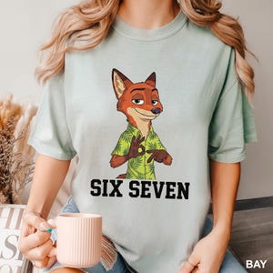 Puede incluir: Camiseta verde claro con un zorro de dibujos animados con una camisa hawaiana verde. El zorro est&aacute; haciendo un gesto con la mano de "OK". El texto "SIX SEVEN" est&aacute; impreso en negrita, en letras negras debajo del zorro.