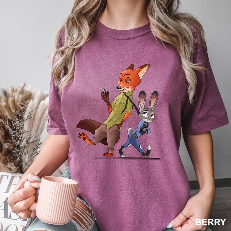 May include: Berry-colored t-shirt featuring a cartoon illustration of Nick Wilde and Judy Hopps from Zootopia. Nick, a fox, is holding a phone, while Judy, a rabbit, appears to be running. The shirt has a relaxed fit.