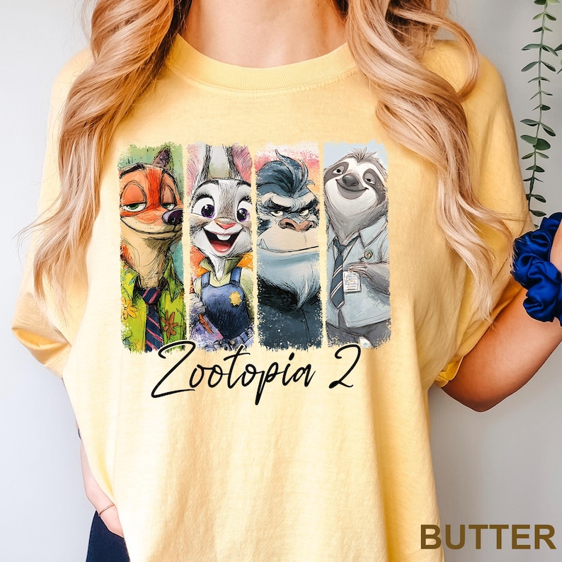 Puede incluir: Camiseta amarilla clara con un estampado colorido de personajes de Zootopia, incluyendo un zorro, un conejo, un gorila y un perezoso. El texto "Zootopia 2" est&aacute; escrito debajo de los personajes. La palabra "BUTTER" est&aacute; impresa en la esquina inferior derecha.