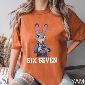Puede incluir: Camiseta naranja quemado con una imagen de dibujos animados de Judy Hopps de Zootopia con uniforme de polic&iacute;a. El texto "SIX SEVEN" est&aacute; impreso en letras blancas debajo. La camiseta es de estilo casual.