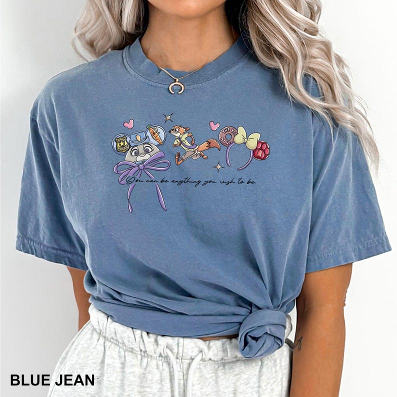 Puede incluir: Camiseta azul vaquero con dise&ntilde;os de personajes de dibujos animados y el texto "You can be anything you wish to be." La camiseta tiene cuello redondo y mangas cortas. La parte inferior de la camiseta est&aacute; anudada.