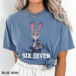 Puede incluir: Camiseta azul vaquero con una imagen de dibujos animados de Judy Hopps de Zootopia. El personaje lleva un uniforme de polic&iacute;a y hace un gesto con la mano. El texto "SIX SEVEN" est&aacute; impreso debajo.