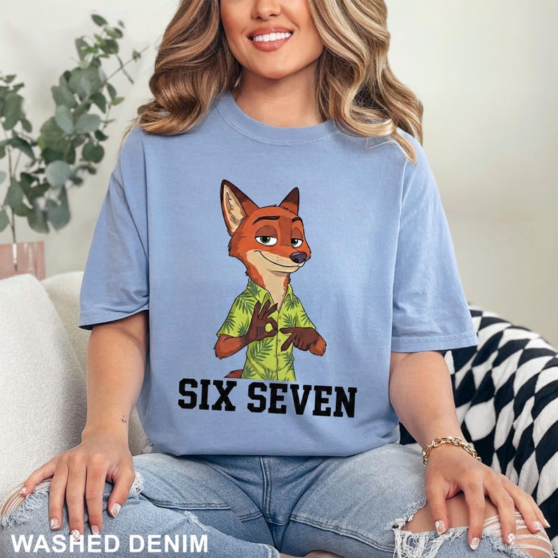 Puede incluir: Camiseta azul claro con un personaje de zorro de dibujos animados con una camisa hawaiana verde. El texto "SIX SEVEN" est&aacute; impreso en negro debajo del personaje. La camiseta est&aacute; etiquetada como "WASHED DENIM" en la parte inferior.