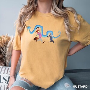 Może przedstawiać: Musztardowa koszulka z kresk&oacute;wkową ilustracją lisa i kr&oacute;lika biegnących z niebieskim wężem. Lis ma na sobie r&oacute;żową koszulę, a kr&oacute;lik granatowy garnitur. Słowo "MUSTARD" jest wydrukowane na dole.