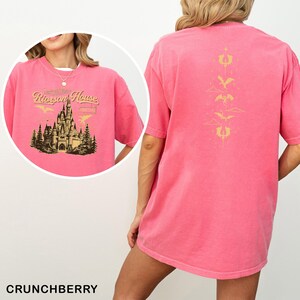 May include: A pink t-shirt with a graphic on the front featuring a castle, trees, and the text "Rixson House". The back of the shirt has a gold design of dragons and clouds. The shirt is a solid color and has short sleeves.