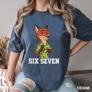 Puede incluir: Una camiseta azul vaquera con un personaje de zorro de dibujos animados con una camisa hawaiana verde. El texto blanco "SIX SEVEN" est&aacute; impreso debajo. La camiseta tiene mangas cortas y cuello redondo.