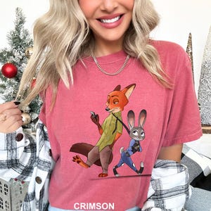 May include: Crimson-colored t-shirt featuring a cartoon illustration of a fox and a rabbit. The fox is holding a phone and the rabbit is wearing a police uniform. The word "CRIMSON" is printed at the bottom.