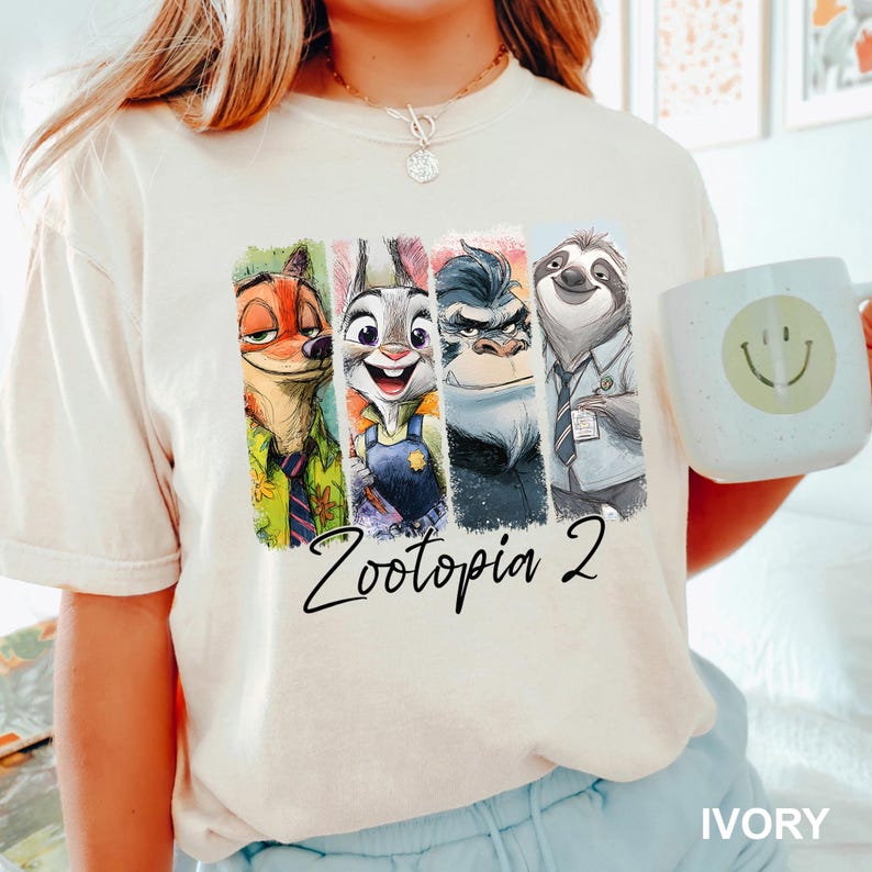 Puede incluir: Camiseta color marfil con un gr&aacute;fico colorido de personajes de Zootopia 2: Nick Wilde, Judy Hopps, Jefe Bogo y Flash. Tambi&eacute;n se ve una taza blanca con una carita sonriente. El texto "Zootopia 2" est&aacute; escrito en negro.