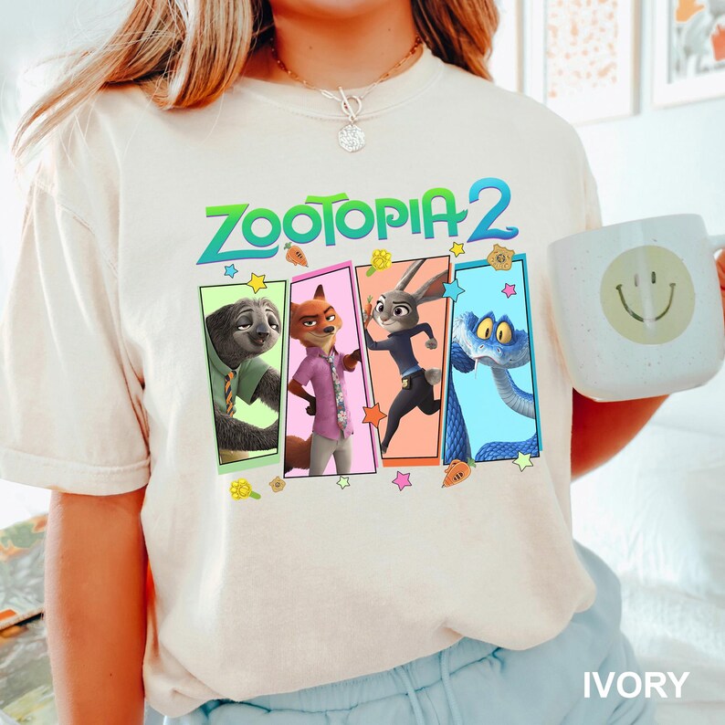 May include: Ivory-colored t-shirt featuring the text "ZOOTOPIA 2" above four character panels. The panels showcase a sloth, fox, rabbit, and snake. A white mug with a smiley face is held in the image.