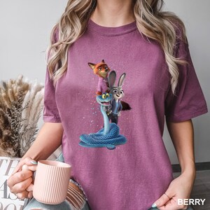 May include: Berry-coloured t-shirt with a vibrant graphic of animated characters. The design features a fox, a rabbit, and a blue snake. The shirt has short sleeves and a crew neck. The word "BERRY" is printed on the bottom right corner of the shirt.