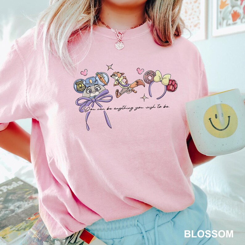 Puede incluir: Sudadera rosa claro con un gr&aacute;fico de personajes de dibujos animados y el texto "You can be anything you wish to be." Una taza blanca con una carita sonriente amarilla se sostiene en la mano.