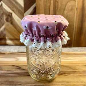 May include: A pink fabric jar lid with a white lace trim and a pattern of bees. The lid is on top of a clear glass jar with the word "Ball" embossed on the side.