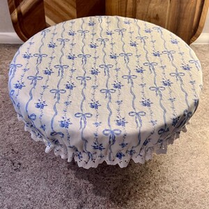 May include: A circular, white fabric bowl cover featuring a blue floral and ribbon design. The cover has a scalloped, lace trim. Wooden chopping boards are in the background.