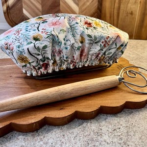May include: A fabric bowl cover featuring a white background with a colorful floral pattern of pink, yellow, and green botanical illustrations, paired with a dough whisk that has a long, light-colored wooden handle and a metal wire loop end.