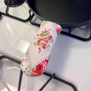 May include: A black cast iron skillet with a fabric handle cover featuring a white background with a pink and red rose floral pattern, green leaves, and a small red bow detail, positioned on a white stovetop with black metal burner grates.