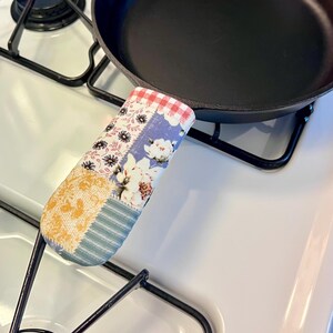 May include: A black cast iron skillet with a fabric handle cover featuring a patchwork design of red and white gingham, white floral patterns on a blue background, yellow lace-like textures, and blue and white striped fabric sections.