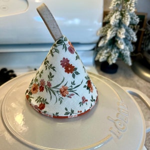 May include: A white ceramic pot with a floral patterned fabric lid. The lid has a loop handle and is shaped like a cone. The pot is sitting on a white countertop.