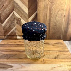 May include: A clear glass jar with a decorative pattern, topped with a navy blue fabric lid. The lid is patterned with white stars and red dots. The jar sits on a wooden cutting board.