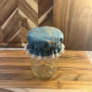 May include: A clear glass Ball jar with a blue fabric lid featuring a floral design and text. The lid is trimmed with white lace. The jar sits on a wooden surface.