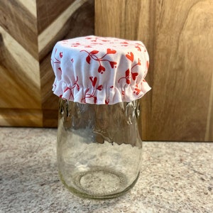 May include: A white fabric jar cover with a red heart and vine pattern. The cover is gathered at the bottom and fits over a glass jar.