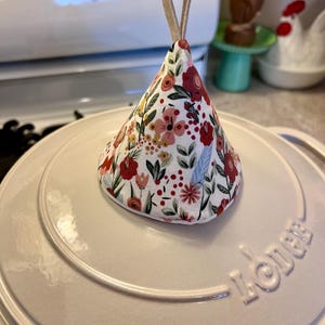 May include: A floral print pot lid lifter with a loop handle. The lifter is cone-shaped and features a white background with red, pink, and yellow flowers and green leaves. It sits atop a white pot lid.