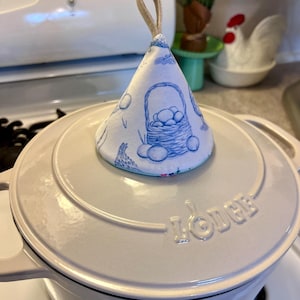 以下が含まれることがあります： 「Lodge」のエンボス加工が施された白い鍋の蓋の上に、円錐形の鍋つかみが乗っています。鍋つかみは、卵と鶏のバスケットの青と白の模様が特徴です。ベージュのループが鍋つかみの最上部に取り付けられています。