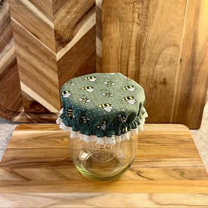 May include: A clear glass jar with a green fabric lid featuring a pattern of yellow and black bees. The lid is trimmed with white lace. The jar is sitting on a wooden cutting board, suggesting kitchen or home decor.