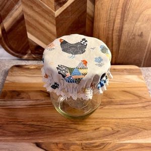 May include: A clear glass jar topped with a fabric cover featuring a pattern of chickens and small flowers. The fabric is beige with black, blue, and orange accents, finished with a white lace trim around the elasticized edge of the cover.