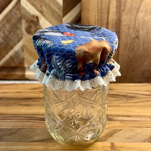 May include: A clear glass Ball jar with a blue fabric lid featuring a farm animal print and white lace trim. The jar is sitting on a wooden surface. The jar is empty.