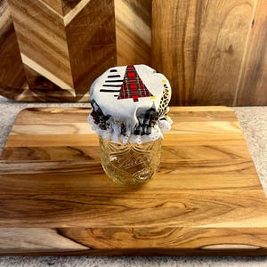 May include: A clear glass Ball mason jar topped with a fabric cover featuring a Christmas tree, leopard print, and black and white buffalo plaid pattern, finished with a white crocheted lace trim around the gathered edge of the fabric.