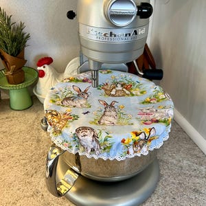 Op de afbeelding: Een zilveren KitchenAid Professional 600 keukenmixer met een blauw-witte komhoes met konijnenmotief. De hoes heeft konijnen, bloemen en vlinders. De hoes heeft een witte kanten rand.