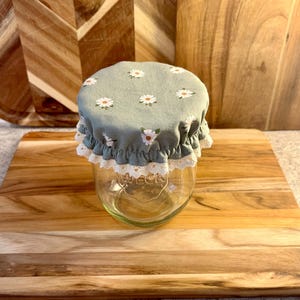 May include: A clear glass jar with a sage green fabric cover featuring a white daisy print. The cover has a ruffled edge trimmed with white lace. The glass jar has the word WECK embossed on the front surface, indicating the brand of the container.