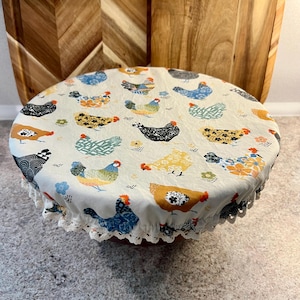 May include: A cream-colored bowl cover with a repeating pattern of colorful chickens. The cover has a white lace trim and is set on a gray surface. Two wooden cutting boards are in the background.