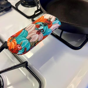 May include: A black cast iron skillet with a fabric handle featuring a colorful pumpkin pattern. The handle is designed to protect hands from heat.