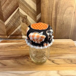 May include: A clear glass Ball jar topped with a fabric lid featuring orange polka dots, orange and white gingham pumpkins, and black and white plaid patterns. The lid has a white lace trim. The jar sits on a wooden surface.