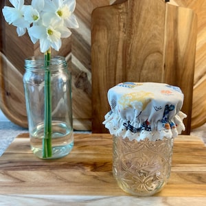 May include: A white fabric jar lid with a floral pattern and lace trim sits on top of a clear glass jar. The jar is on a wooden cutting board with a vase of white daffodils.