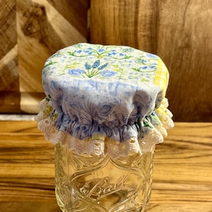 May include: A blue and white floral fabric jar lid cover with white lace trim. The lid cover is on top of a clear glass jar with the word "Ball" embossed on the side.